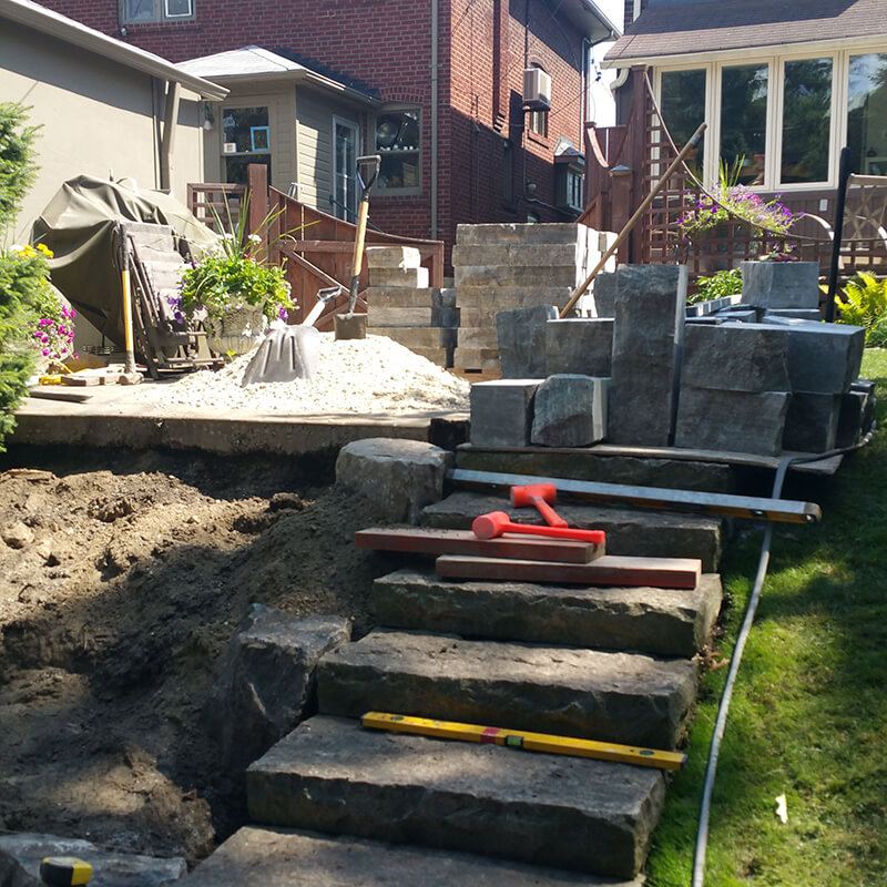 Armour stone steps being set in a backyard, with levels and mallets on the treads and stacks of cut stone behind