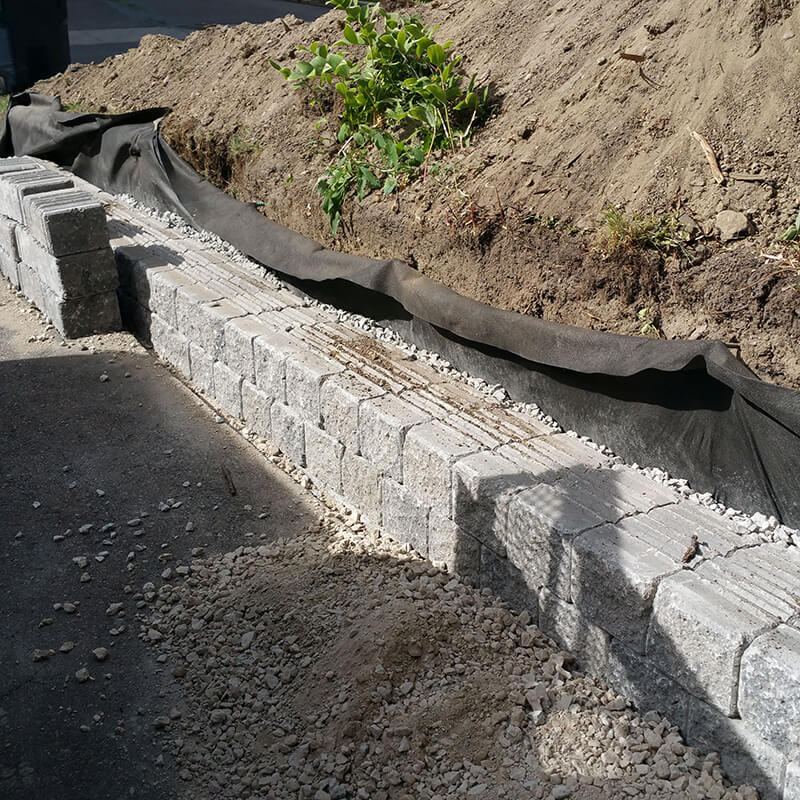 Grey precast block courses going in along an asphalt driveway, with black filter fabric against the soil behind