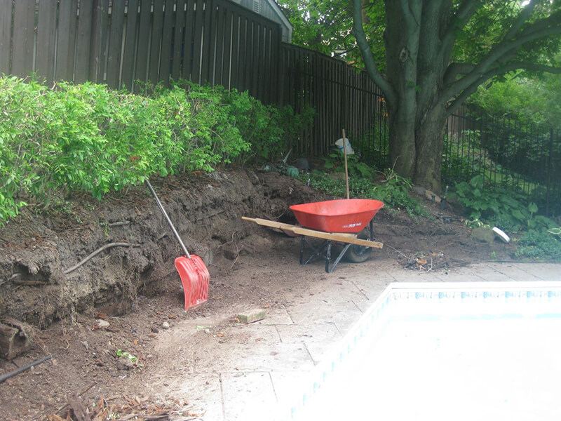 Bank beside the pool cut back by hand after the old railroad-tie wall came out, with a wheelbarrow and shovel on the deck