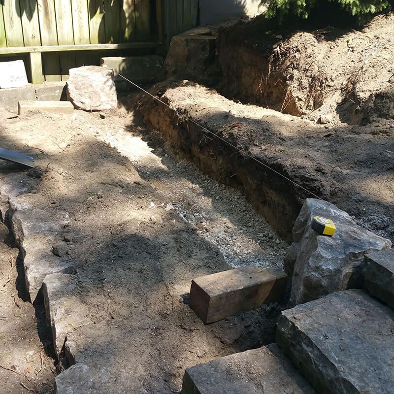 Excavated trench with a gravel base and a string line beside rough armour stone steps, with a timber fence behind