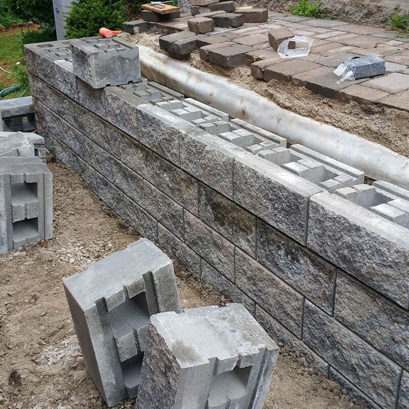 Large hollow split-face precast blocks stacked on a wall, with a white drainage pipe behind and spare blocks on the ground