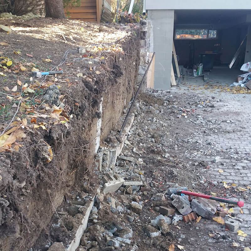 Old brick-and-block retaining wall broken out beside a garage and interlock driveway, with rubble on the pavers and the soil cut back behind