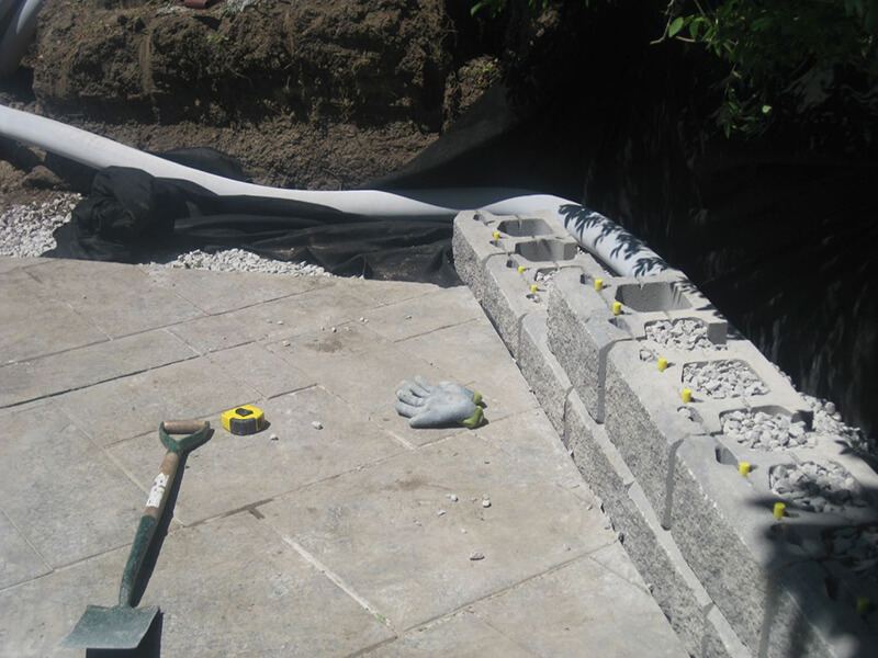 First courses of hollow precast blocks filled with clear stone and pinned, set by hand on the edge of a patterned concrete pool deck, with drain pipe and filter fabric behind