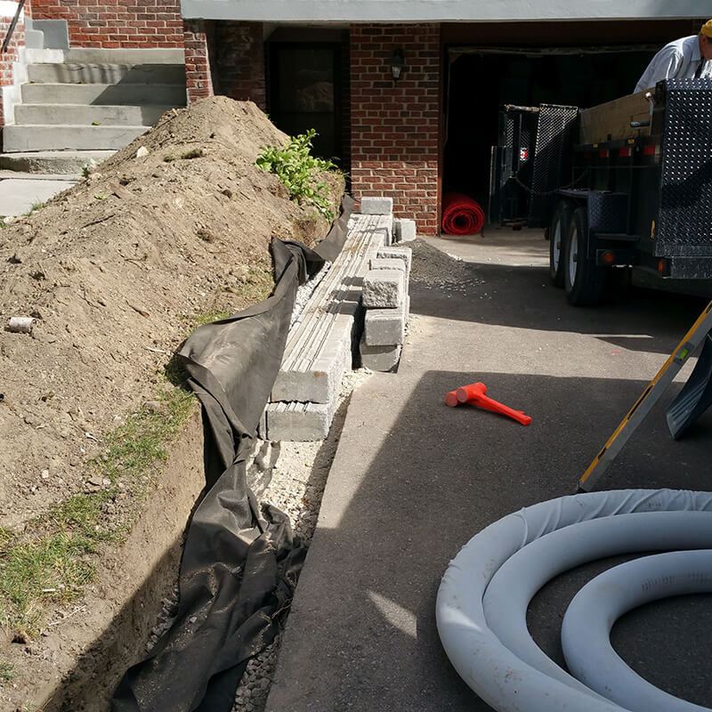Excavation along a driveway beside a brick house, lined with filter fabric, with the first blocks set and a coil of socked weeping tile on the driveway