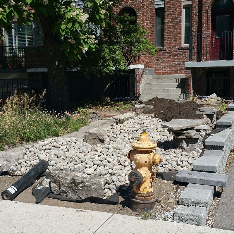 Front yard mid-project: river rock and armour stone being placed around a fire hydrant, with coping blocks stacked beside the new wall and a roll of filter fabric