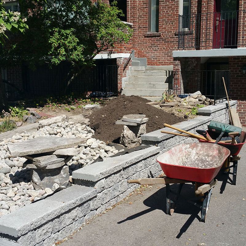 Finished block wall with coping along the driveway, river rock, stone slabs and a pile of soil behind it, wheelbarrows in front