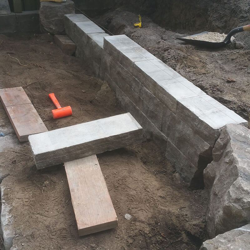 Dry-stacked Eramosa wall courses ending at a large armour stone bookend, with a long natural stone coping piece laid out on boards in front
