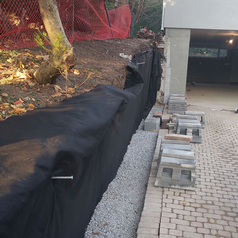 Black filter fabric pinned over the excavated slope above a bed of clear stone, with precast blocks stacked on the driveway