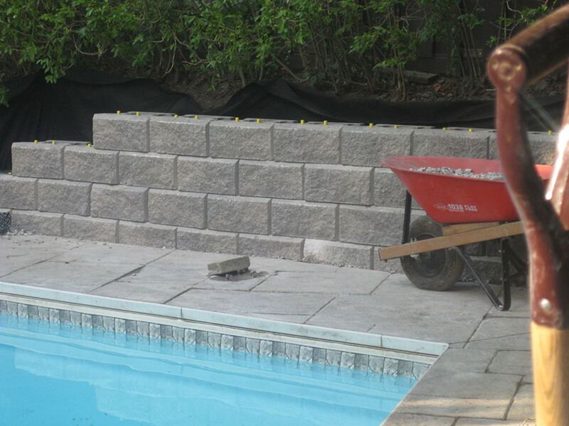 Split-face precast block wall going up beside a swimming pool, with yellow pins on the top course and a red wheelbarrow of gravel on the pool deck