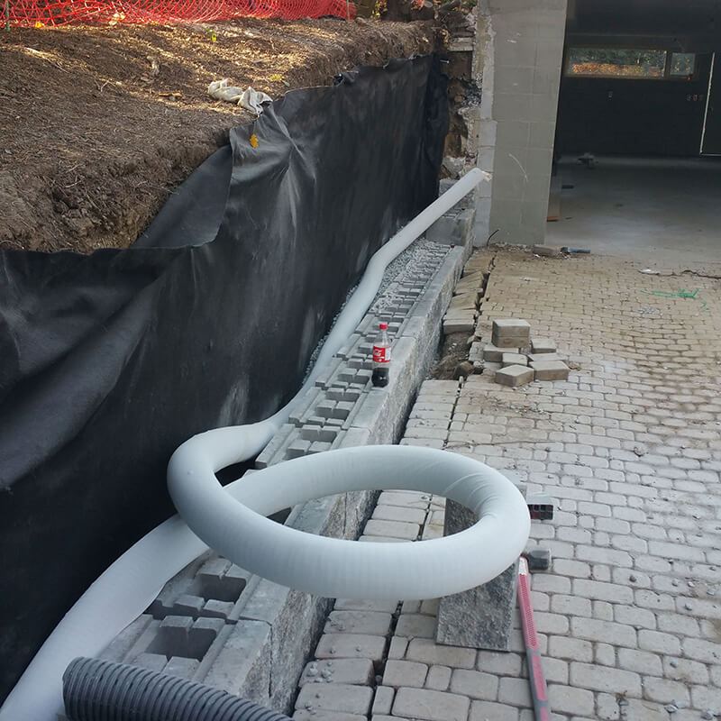 First course of hollow block on its base, with black filter fabric lining the slope and a coil of white socked drain pipe beside an interlock driveway
