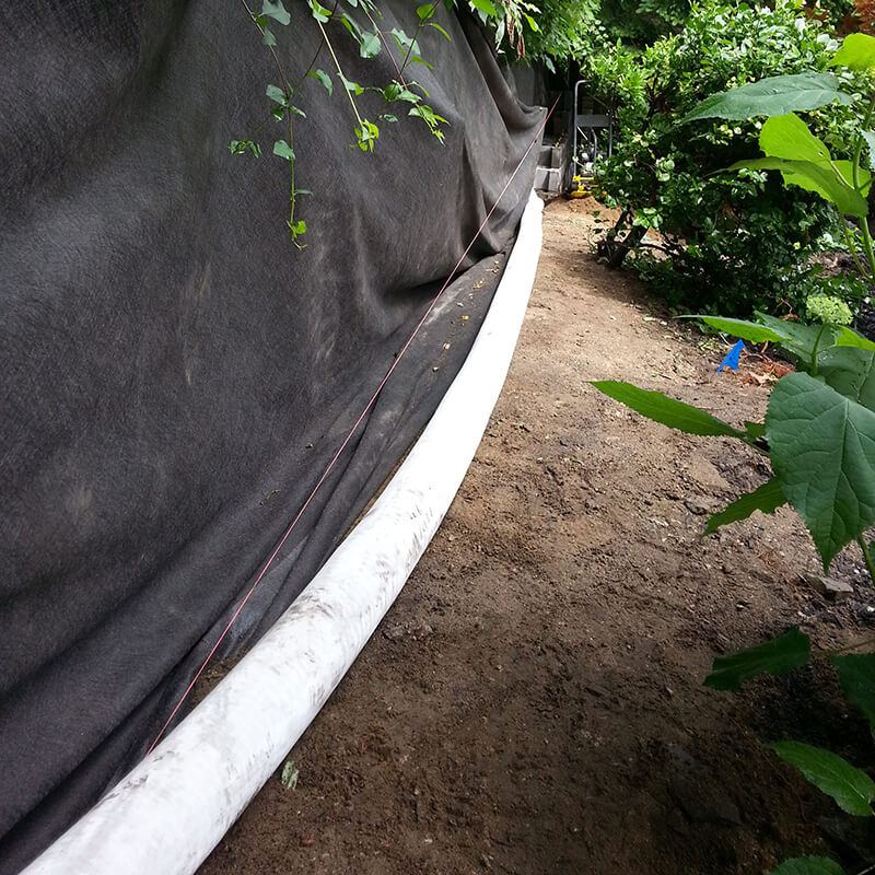 Black filter fabric lining an excavated bank behind a curving wall, with a white socked drain pipe along its base and garden plants alongside