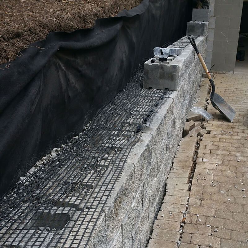 Geo-grid mesh laid over a course of split-face precast block, with black filter fabric against the soil behind and a shovel on the pavers