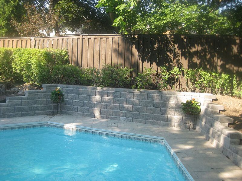 Backyard pool edged by a grey precast block retaining wall, with a tall wooden privacy fence along the top of the yard