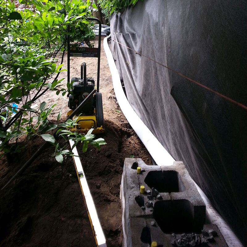 Hollow precast block with yellow fibreglass pins in a trench, white drain pipe along filter fabric on the bank, and a plate compactor behind