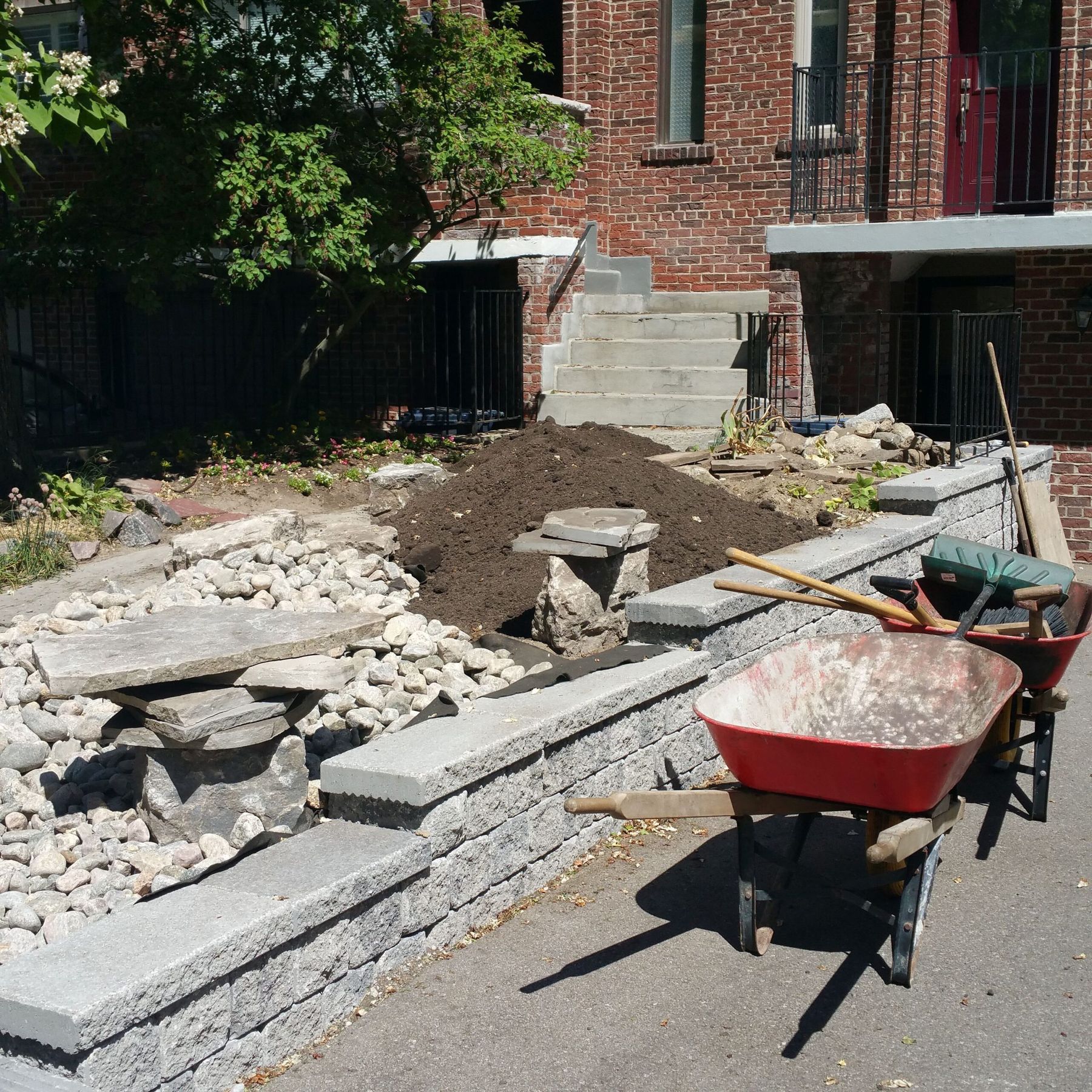 Small precast block retaining wall with grey coping along a driveway, mid-build, with a wheelbarrow, a pile of topsoil and river rock in front of a brick house