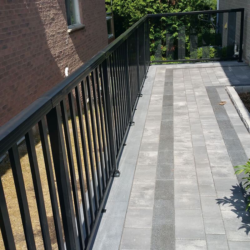 Grey paver walkway with a dark inlaid border running along the top of a retaining wall, edged by a black metal railing beside a brick house