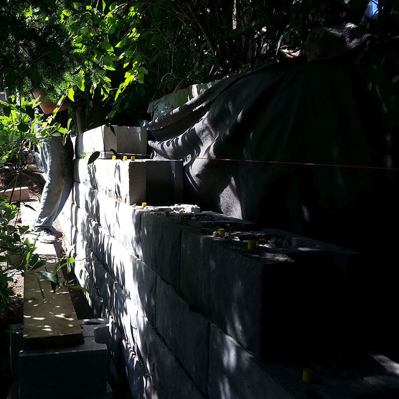 Hollow precast block courses with yellow pins rising against black filter fabric in a shaded garden