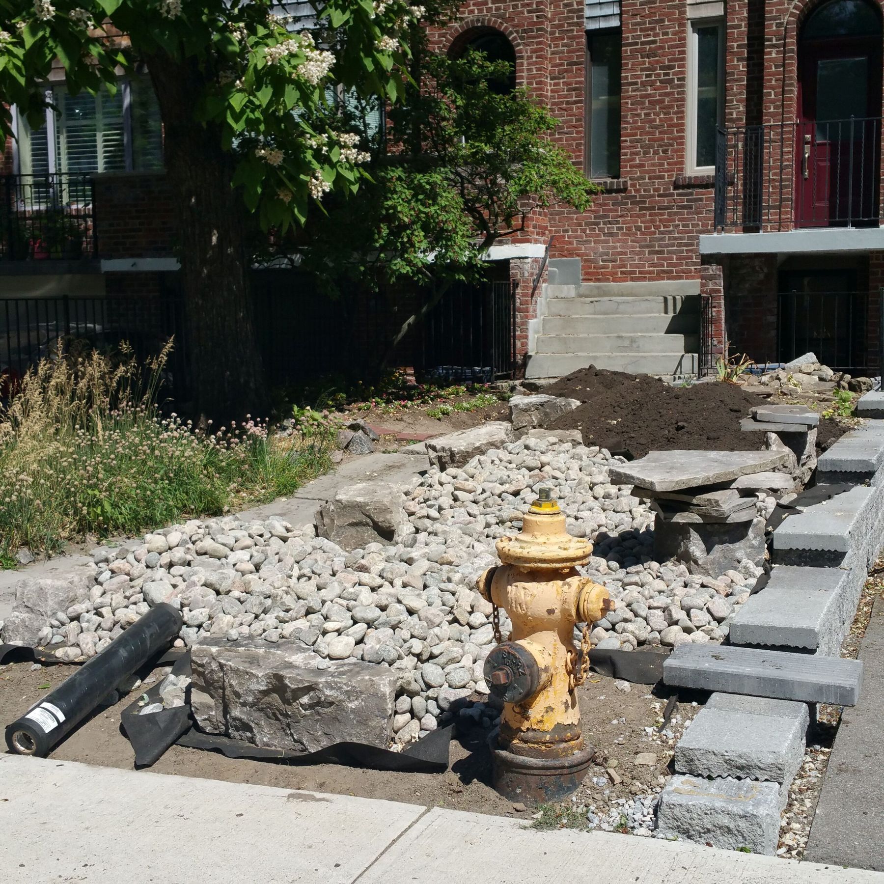Front-yard landscaping taking shape beside a red-brick house: river rock and armour stone around a yellow fire hydrant, grey precast block caps along the walk, a pile of topsoil and a roll of landscape fabric