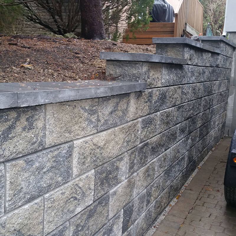 Tall grey split-face precast block retaining wall with stepped caps beside an interlock side path