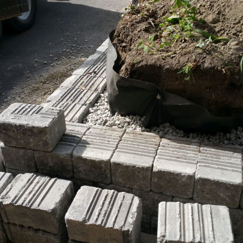 Grooved precast blocks set on a clear gravel base, with black filter fabric wrapping the soil behind the first course