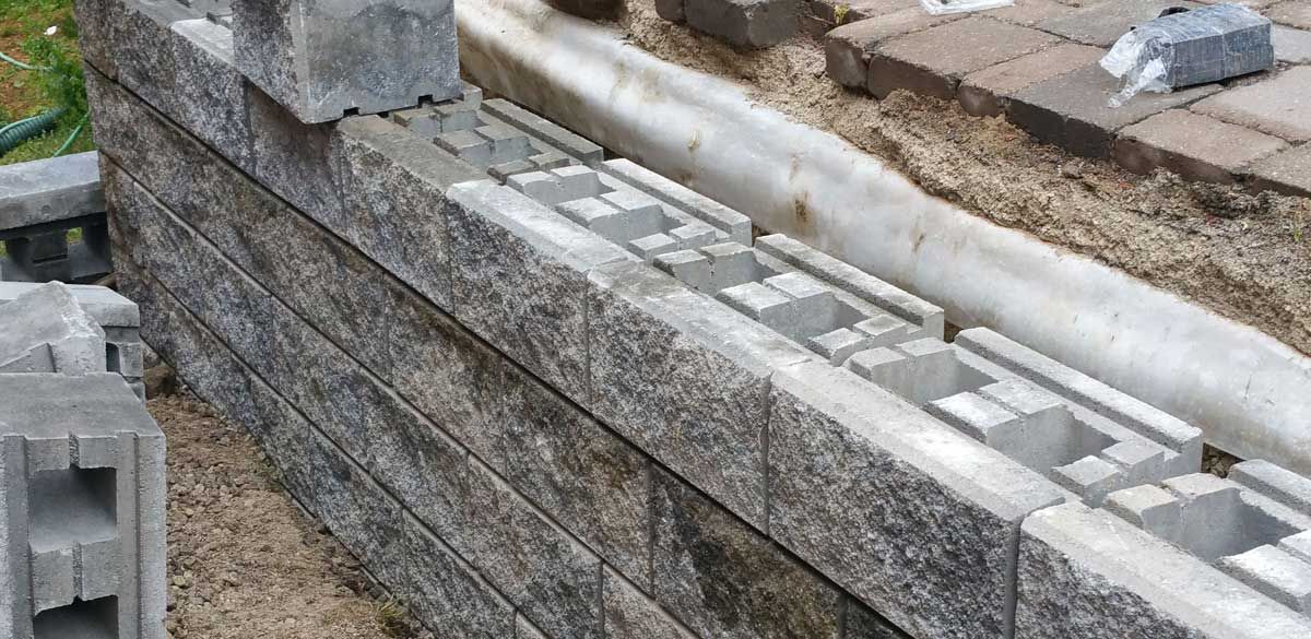 Grey split-face precast retaining wall blocks being stacked mid-build, with open hollow cores and a white drainage pipe laid behind the courses below interlock pavers
