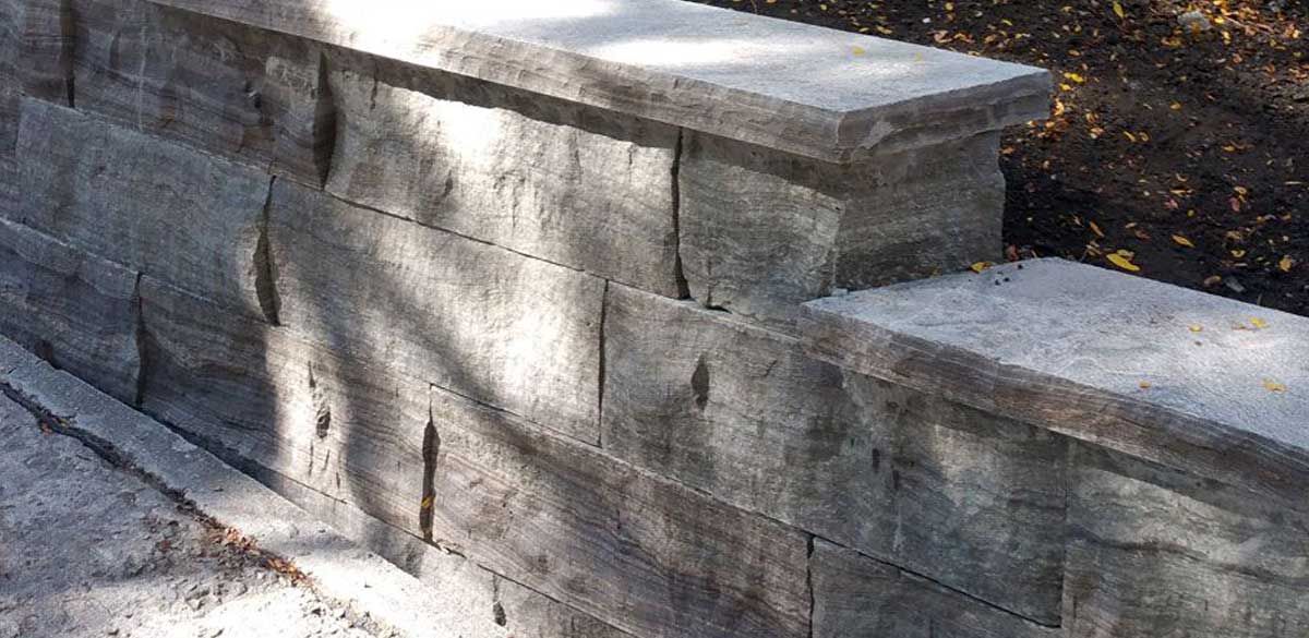 Dry-stacked grey natural stone retaining wall with thick stone coping stepping up beside a path, with fallen leaves on the garden bed above